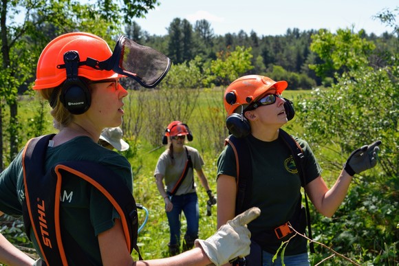 A group of people wearing work clothes, orange hardhats, gloves and other safety equipment stand outside on a sunny summer day.