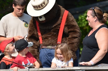 A costumed Smokey Bear impersonator leans forward while talking to smiling, young girls and boys near a fence