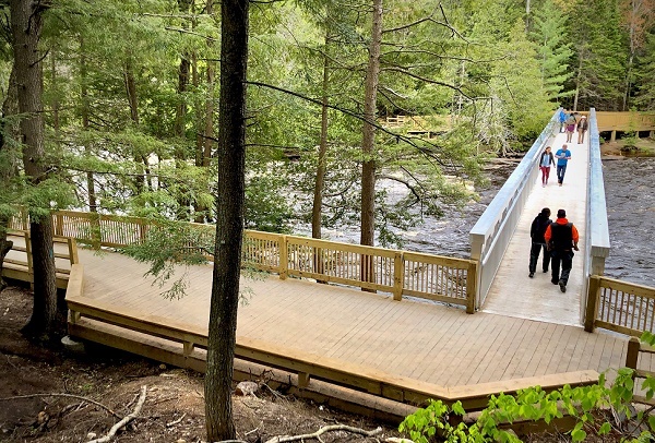 Small clusters of people walk across a new aluminum bridge set over dark, foamy rapids. Large wooden platform in foreground amid forest.