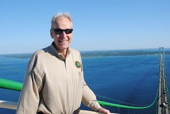 Smiling, older, white-haired man wearing black sunglasses and long-sleeved khaki shirt with DNR logo, atop a tall, green bridge; blue water all around