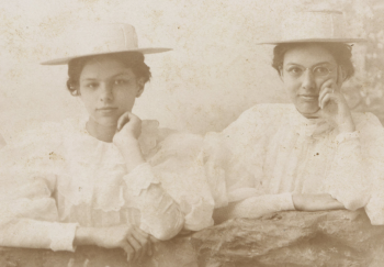 Two women leaning on a rail, both wearing white blouses with big puff sleeves, fabric covered summer hats