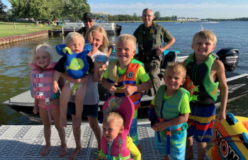 Two conservation officers stand, smiling, behind a large group of life-jacketed children on a dock.