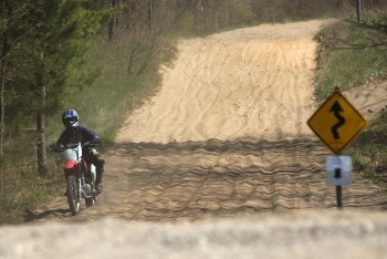 A person riding a dirt bike drives down a sandy slope; in the foreground, a road sign indicating a twisting path is slightly blurred.