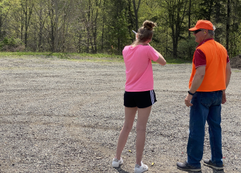 An older man wearing blaze orange stands next to a young woman aiming a rifle.