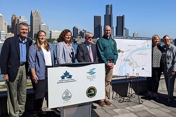 Several men and women in business casual stand around podium, next to a map of Michigan and Ontario, river skyline behind them