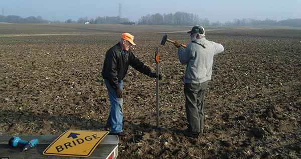 two men pound a post with an orange snowmobile trail marker into the ground
