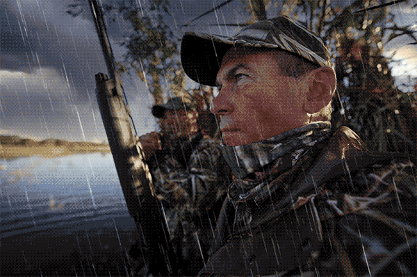 A pair of duck hunters wearing camouflage clothing sitting in a wetland with dark clouds in the sky and animated rain falling.