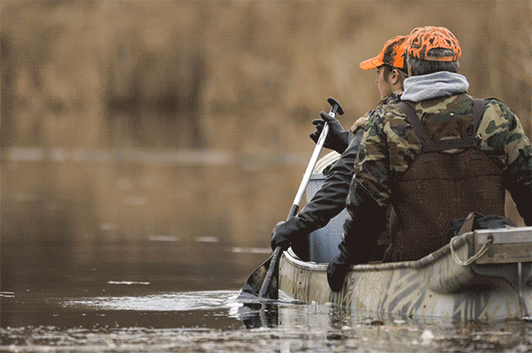 Two trappers wearing camouflage in a canoe paddle through a wetland with animated waves around the canoe.