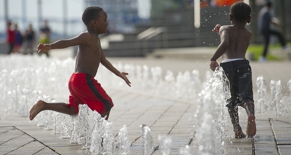 two little boys, one in red swim trunks and the other in navy blue swim trunks, run through water spray of a cement splash pad in a city area