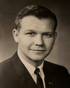 A black and white photo of a man with pale skin and close-cropped hair wearing a formal suit. He has a slight smile on his face.