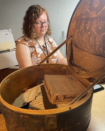 Jessica Bigelow, a young person with pale skin and shoulder-length curly blond hair, inspects an old hat box full of artifacts.