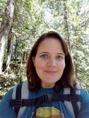 Smiling photo of Andrea Stay with a backpack on. She is a caucasian woman with brown hair with trees in the background. 
