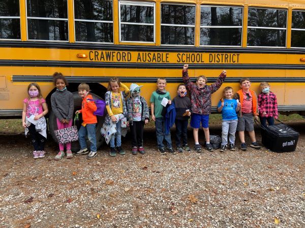 Children stand smiling outside a school bus on their way to a field trip in the forest.