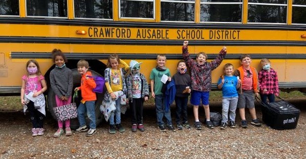 Children stand in front of a yellow school bus in a forest setting. 