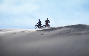 Two adults in full biking gear stand on the precipice of a wind-swept, silvery sand dune.