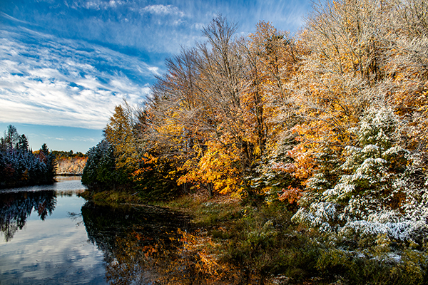 The changing of the seasons is shown with snow-covered trees and blue skies above a lake.