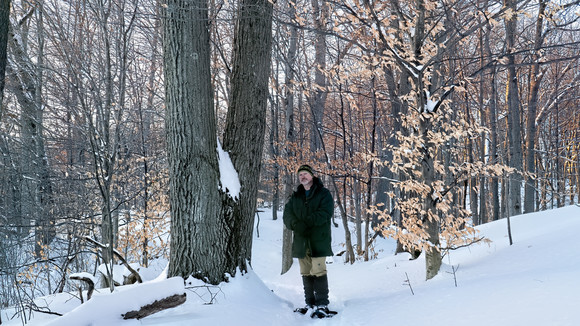 A man in snow gear and wearing snowshoes stands in a snow covered forest to the right of a large tree.