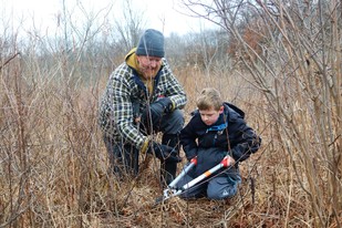 A man and a young boy kneel in dried grasses and shrubs and cut stems with a pruning tool.