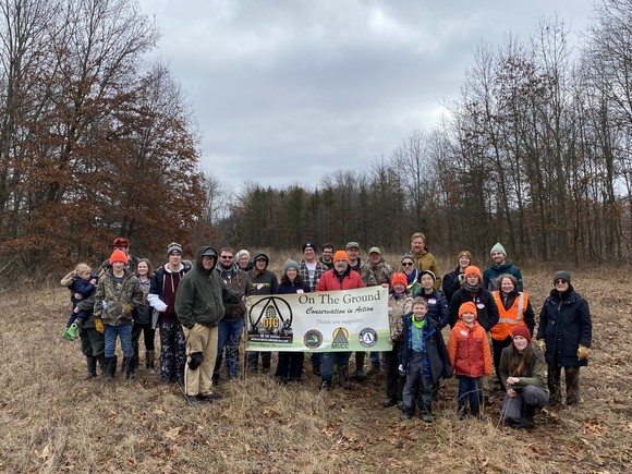 A group of people wearing outdoor gear stand in a forest opening holding a banner for the On The Ground program.