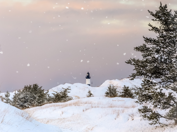 The top of a lighthouse peeks out from behind snow-covered dunes as a sherbert-pink sky drops large, gentle snowflakes.