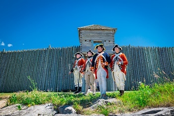 A group of four adult men with pale skin stand in front of a historic fort dressed in historical soldiers' garb, smiling faintly into the distance.