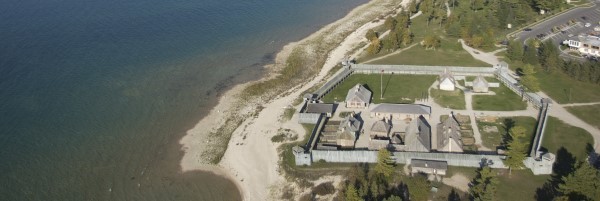 A historic wooden fort sits on the calm shoreline of Lake Michigan.