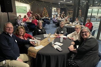About two dozen smiling men and women seated at round tables in a glass-walled atrium, strings of white sparkle lights on back wall