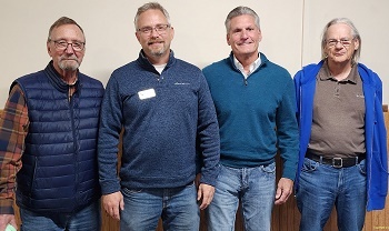 Four smiling, older men with salt and pepper hair, three in glasses, wearing blue jeans and various blue pullover fleeces and flannel