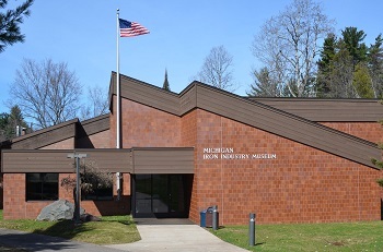 entrance view of a cocoa-colored brick building with angular, dark brown roof lines, white lettered Michigan Iron Industry Museum on front