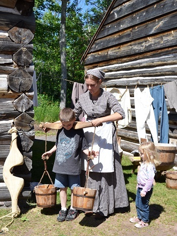 A woman dressed in a black-and-white checked, old-fashioned dress and kerchief places a wooden slat with two roped buckets on a young boy's shoulders