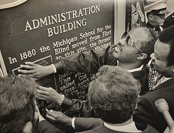 Sepia-toned photo shows two Black men, one wearing glasses, touching the Braille part of a sign labeled Administration Building, as others look on
