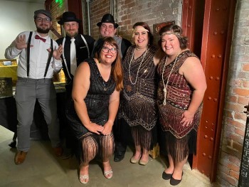 A group of people in flapper dresses and historically-styled trousers, suspenders and bow-ties smile at the camera.