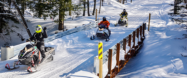 Three snowmobilers crossing trail bridge, one snowmobiler waving