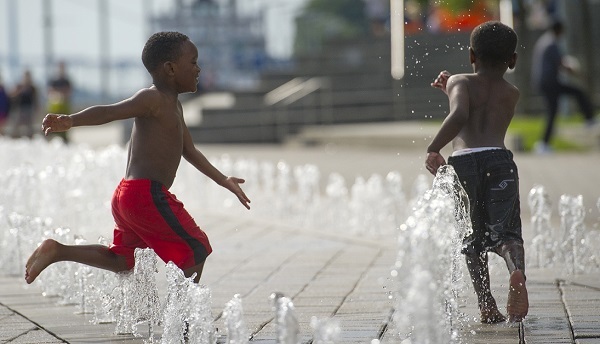 two little boys, one in red swim trunks and the other in blue, laugh and chase each other through small fountains of water on a concrete splash pad