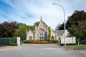 street entrance at the opening in a dark green vertical fence, a sign reading Elmwood Cemetery, and a ornate tan and cream building