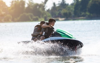 Two people wearing life jackets ride a jetski across a calm summer lake.