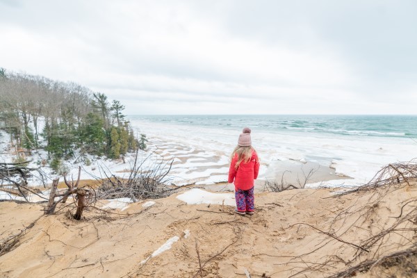 A small child decked in pink winter gear stands on a bluff overlooking a wintry Great Lakes shoreline.