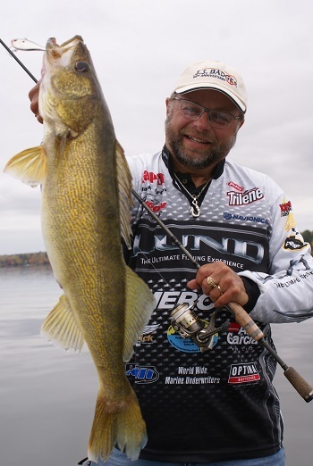 A neatly bearded, smiling man wearing an S.S. Badger ballcap and a white, logoed, long-sleeved shirt holds a fishing rod and a large walleye