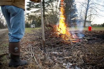 A burning brush pile is supervised by an onlooker holding a shovel.