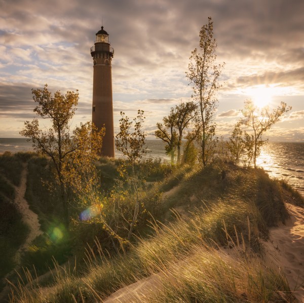 A hazy sunset casts golden light on a grassy lakeshore dune, the spire of a lighthouse jutting from the sand.
