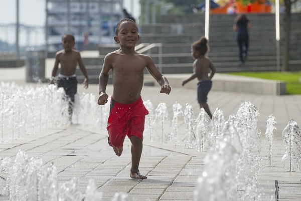 a smiling young boy in navy blue swim trunks jumps in the spray of a concrete splashpad, with two small boys playing behind him