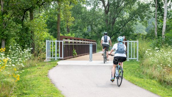 Two cyclists enjoy a paved bike path lined with trees and wildflowers