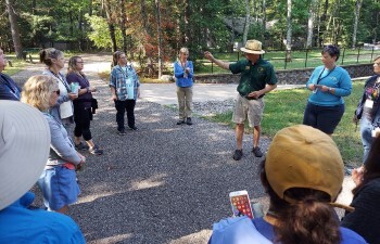 A group of people stand in a circle listening to a facilitator talk about birds.