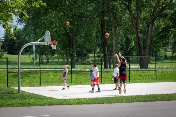 A group of adults play basketball on a newly constructed court as a gentle summer sun shines down on them.