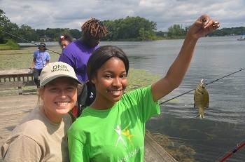 On the beach, a young girl in a bright green shirt holds up a fish she caught as a young woman in a DNR ballcap looks on