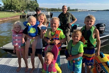 a small group of very young, smiling girls and boys in bathing suits and life jackets stand on a pier, a conservation officer is behind them