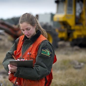 A forester in front of heavy equipment looks at sheets on a clipboard