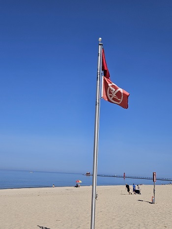 Two red flags hang from a tall pole on a sandy beach, near the shoreline. Double red flags on Great Lakes beaches mean don't go in the water.