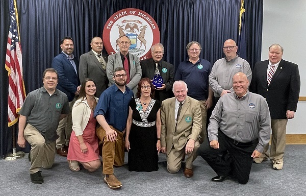 An older man, back row center, holds a blue glass award, surrounded by a dozen smiling men and women, in front of State of Michigan seal