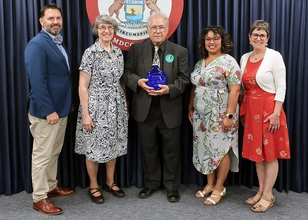 An older man, center, holds a blue glass vase, flanked by a man and three women, in front of Michigan state seal. Everyone is dressed nicely.